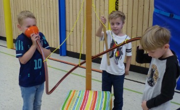 Drei Jungen im Grundschulalter experimentieren in einer Turnhalle an einer MINIPHÄNOMENTA-Station mit einem orangefarbenen Trichter und Schläuchen, um Schallwellen und Töne zu erforschen.