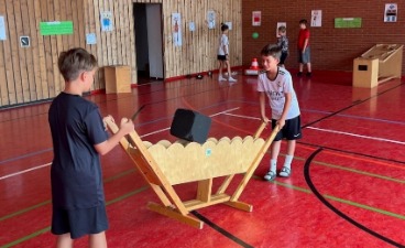 Zwei Grundschulkinder experimentieren in einer Turnhalle gemeinsam an einer hölzernen MINIPHÄNOMENTA-Station mit einem großen, schwarzen Würfel.