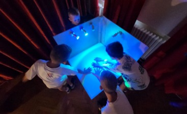 Fünf Kinder betrachten fasziniert ihre Hände in einem weißen Gehäuse, das von innen hellblau beleuchtet wird. Die Kinder stehen dicht gedrängt und blicken von oben in das Gehäuse, während drei Lichter das Innere erhellen.