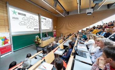 Teilnehmende einer Fachtagung sitzen in einem Hörsaal und verfolgen einen Vortrag mit Präsentation auf zwei großen Leinwänden.