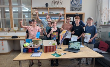 Neun lächelnde Jugendliche und Betreuer der Bildungsinitiative stehen im Werkraum Augsburg hinter einem Tisch und zeigen den Daumen nach oben. Auf dem Tisch sind gebastelte Elektronik-Projekte wie eine Station mit der Aufschrift „Aurora“, ein Laptop mit einem Programmier-Code sowie Urkunden und eine kleine code your way-Fahne zu sehen.