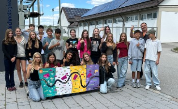 Eine Gruppe von Schülerinnen und Schülern posiert lächelnd mit Daumen nach oben vor dem LEW Technologiezentrum und hält ein buntes „code your way“-Plakat.  