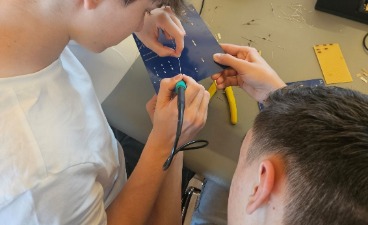 Zwei Jugendliche arbeiten gemeinsam mit einem Lötkolben an den Kontakten auf der Rückseite einer blauen Platine.
