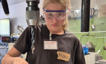 Ein Mädchen mit Schutzbrille konzentriert sich beim Bedienen einer Ständerbohrmaschine in einer Werkstatt.