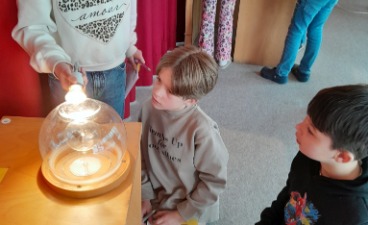 Drei Grundschulkinder knien und stehen an einer hölzernen Experimentierstation und beobachten aufmerksam eine leuchtende Glühbirne unter einer Glaskuppel. 