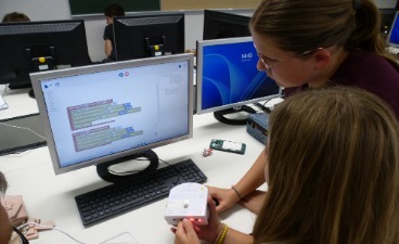 Zwei Schülerinnen programmieren an einem Computerbildschirm einen weißen Thymio-Roboter.