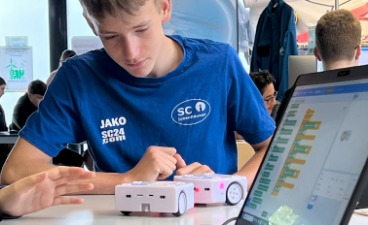 Ein Schüler beobachtet zwei ferngesteuerte Objekte auf einem Tisch. Im Vordergrund steht ein Laptop, über den sie gesteuert werden.